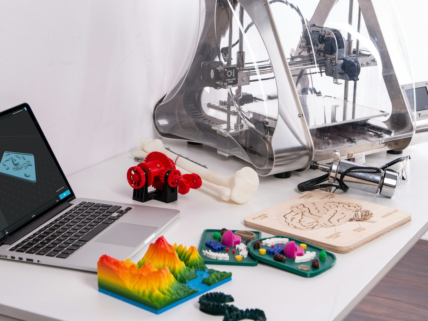 Projeto no computador ao lado da impressora 3D e de peças impressas